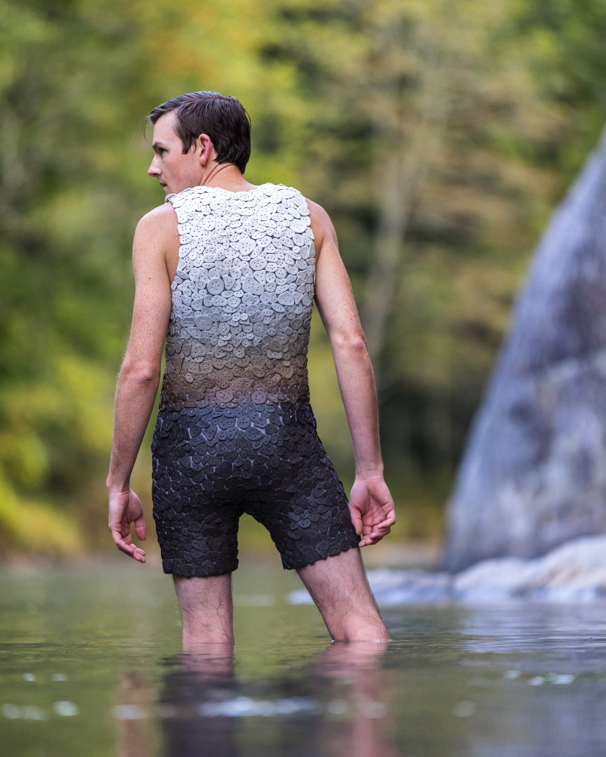Shae Bishop stands in knee-deep water with his back to the viewer while wearing a one-piece suit made with tiles