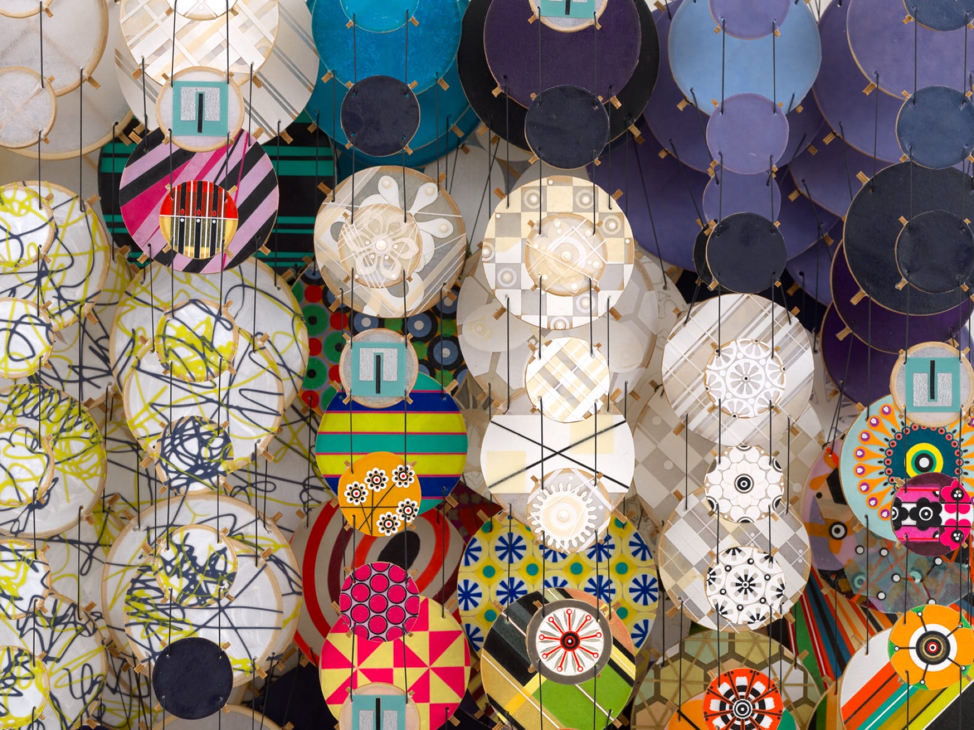 a detail of an abstract artwork by Jacob Hashimoto made from twine and numerous colorful, patterned paper discs installed in rows for a layered effect