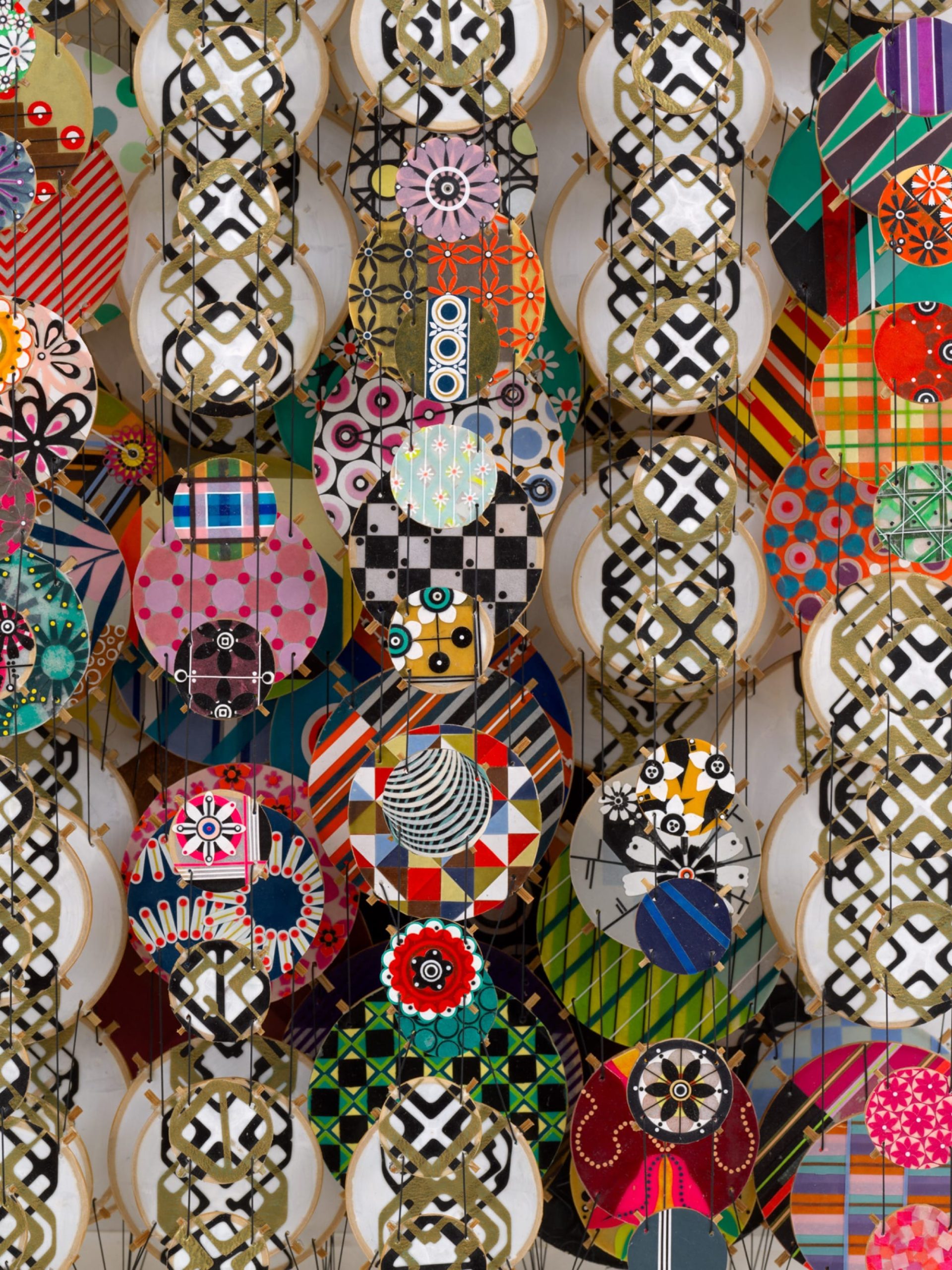 a detail of an abstract artwork by Jacob Hashimoto made from twine and numerous colorful, patterned paper discs installed in rows for a layered effect