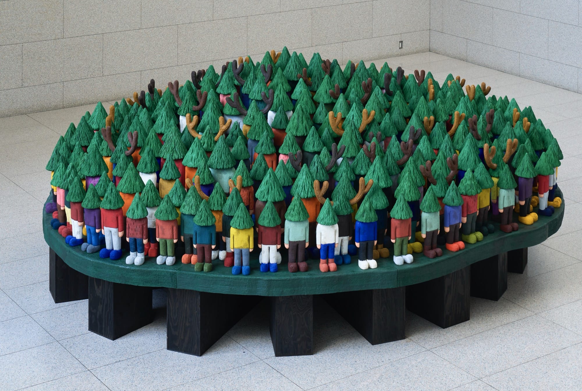 a sculptural installation by Yuichi Hirako of an oversized table covered with cartoonish figures with evergreen trees for heads