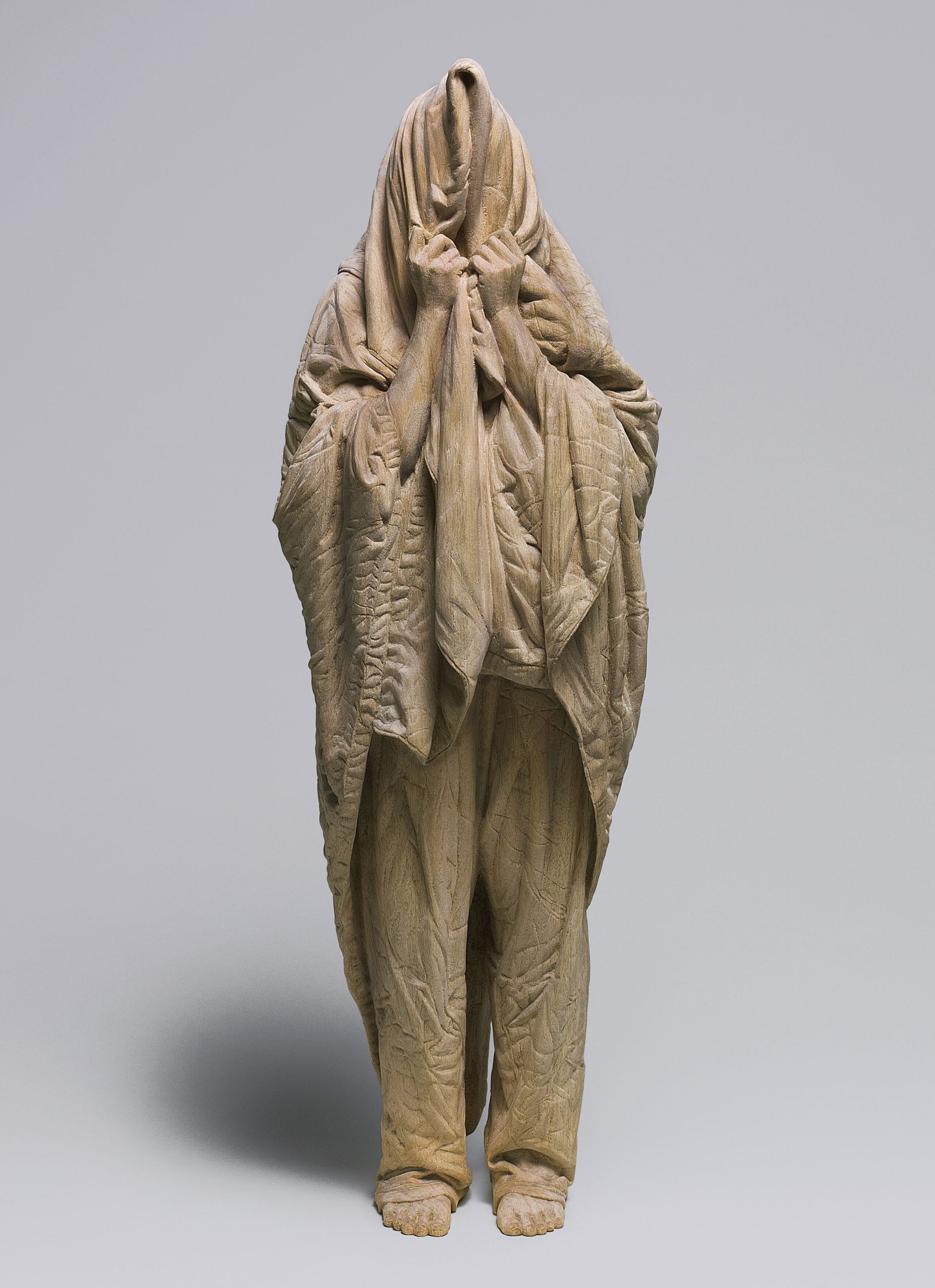 a carved wooden sculpture by Ikuo Inada of a figure standing with a blanket over their head as if very sleepy